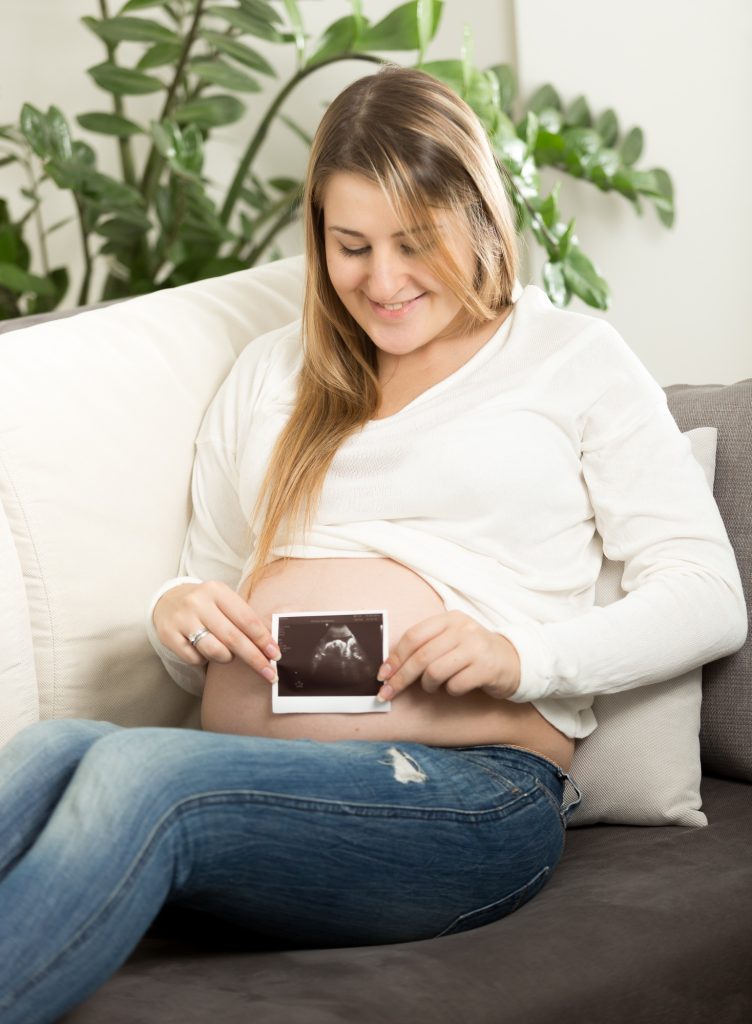 smiling pregnant woman sitting on sofa and holding ultrasound fetus scan